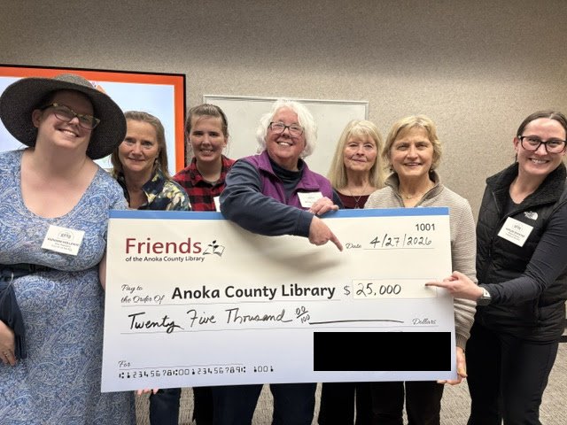 Seven white women from the Friends' Board stand behind a large prop check dated 04/27/2026 and made out to Anoka County Library for the sum of $25,000. A couple point proudly at the total box.