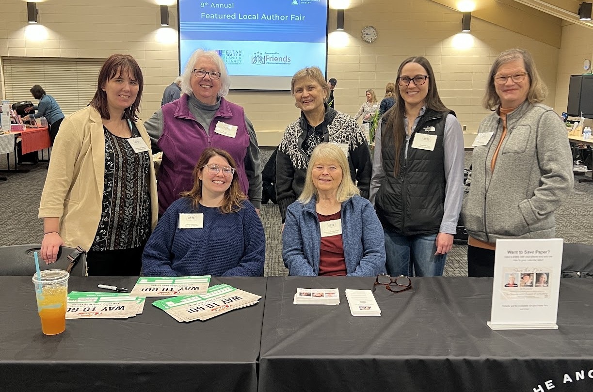 Seven white women from the Friends' Board are behind a black-clothed table smiling at the camera. Behind them a large screen gives the event title, '9th Annual Featured Local Authors Fair,' and logos of the sponsors: Clean Water Land and Legacy Amendment and Friends of the Anoka County Library.