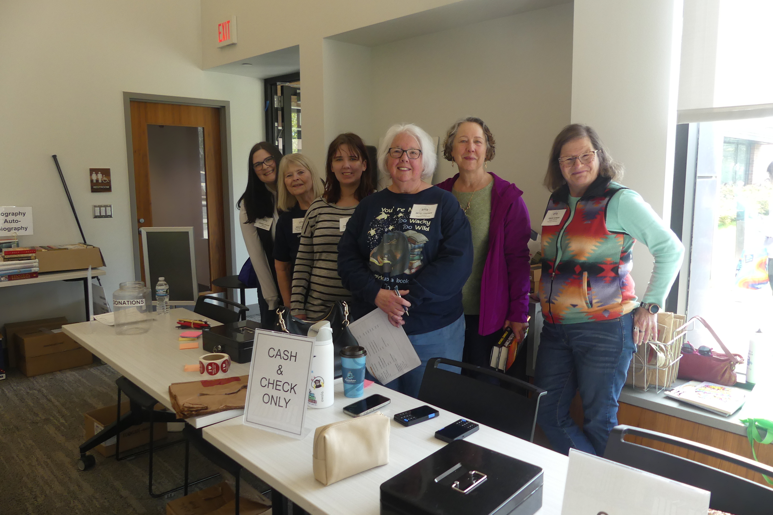 Six Friends volunteers stand behind a check-in table with name tags, a donation jar, and a "Cash & Check Only" sign during a book sale.